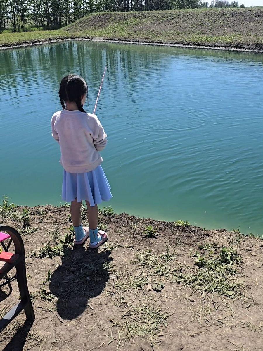 A girl fishing from the bank, the pond stretching out ahead