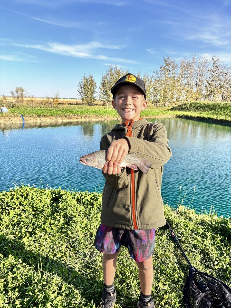 A boy holding up the trout he caught, pond behind him